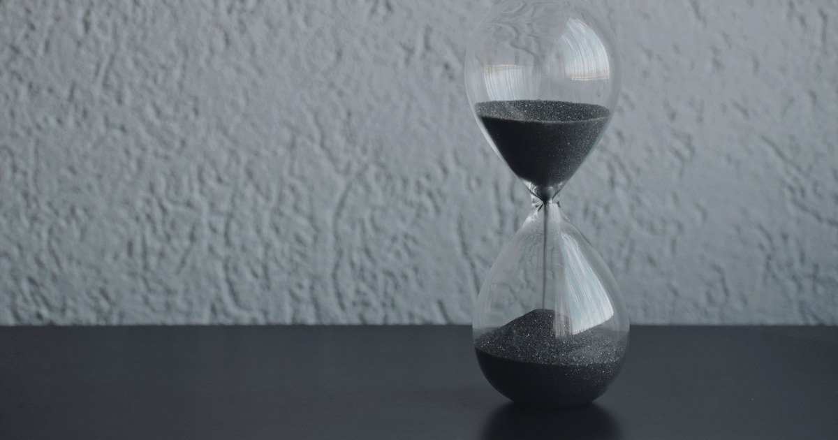 An hourglass with dark sand standing on a table against a gray wall